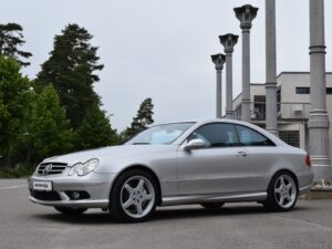 A silver two-door Mercedes-Benz sports car parked in front of a modern building with tall columns and surrounded by trees on a cloudy day.