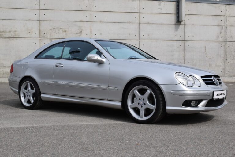 A silver two-door Mercedes-Benz coupe is parked on a paved surface in front of a concrete wall.