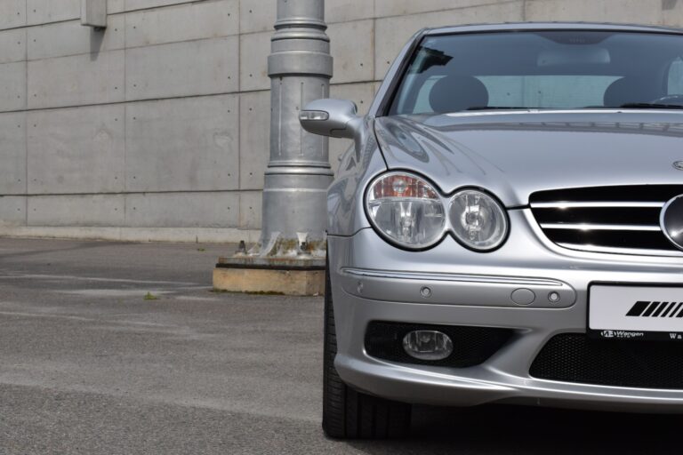 Close-up of the front right side of a silver car parked next to a concrete wall and a large metal post. The image highlights the headlights, grille, and part of the windshield.