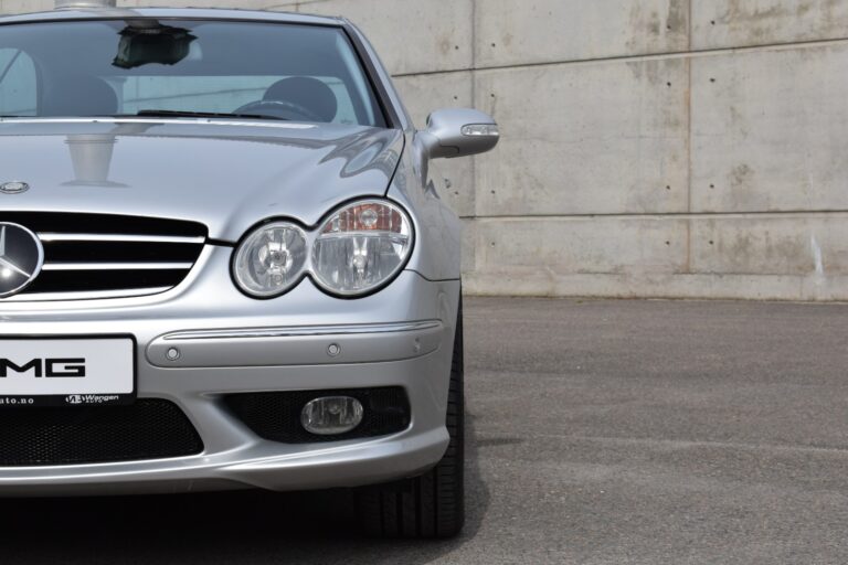 A silver AMG Mercedes-Benz car is parked on a gray asphalt surface, with a concrete wall in the background. Image shows the front-left side of the vehicle.