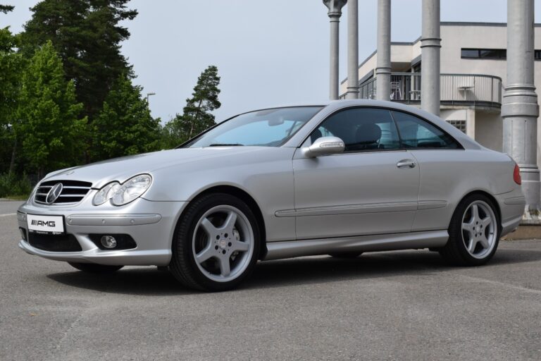 A silver Mercedes-Benz CLK coupe is parked on a paved surface, with a modern building and trees in the background.