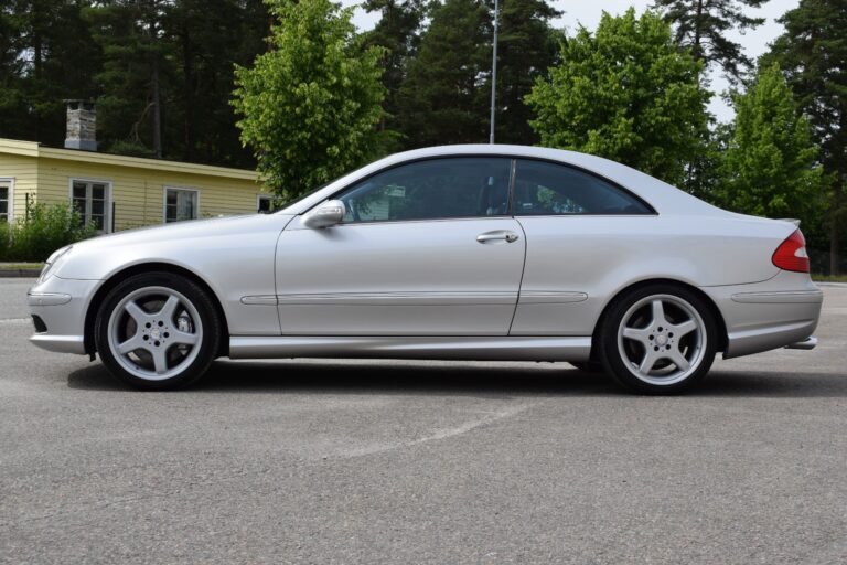 A silver two-door coupe car is parked on a paved surface with a yellow building and trees in the background.