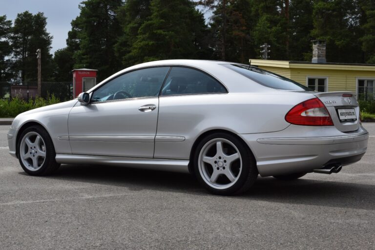 Silver two-door car parked on a paved surface with trees and a yellow building in the background.