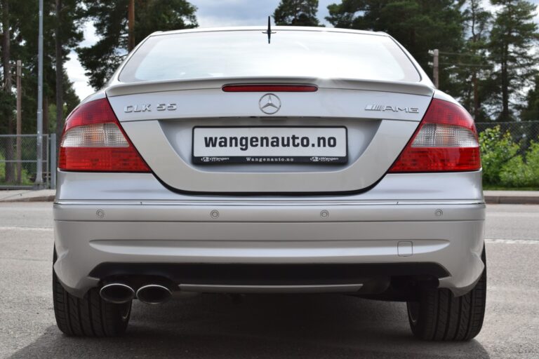 Rear view of a silver Mercedes-Benz CLK 55 AMG parked outdoors, showcasing the car's rear design, dual exhausts, and license plate with the text "wangenauto.no". Trees and a fence are in the background.