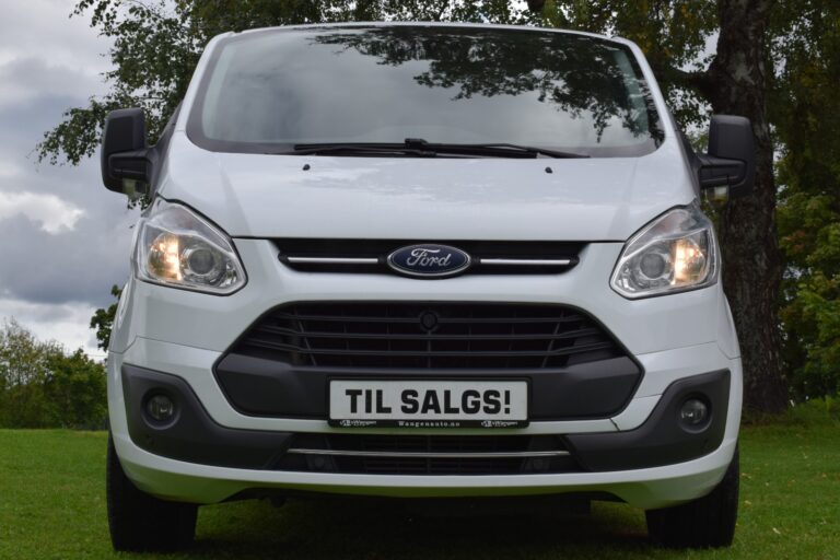 A white Ford Transit van with a "Til Salgs!" sign on the front grille, parked on grass with trees in the background.