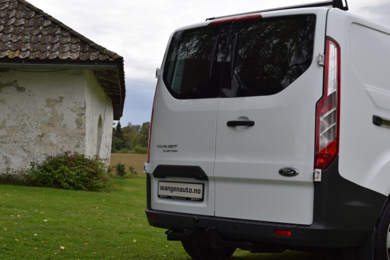 A white Ford Transit Custom van is parked on a lawn near an old building with peeling paint. "wangeanuto.no" is written on the van's license plate.