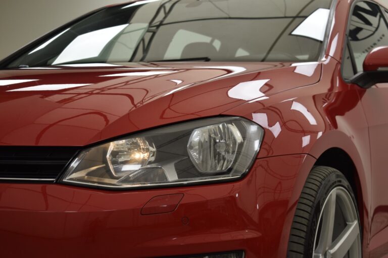 A close-up of the front left side of a red car, focusing on the headlight and part of the hood and bumper. The car is indoors with reflections visible on its surface.