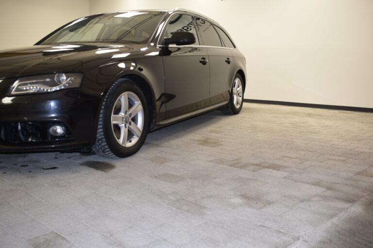 Black sedan parked indoors on a tiled floor, with partial view focusing on the front left side of the vehicle. The garage appears well-lit with plain, light-colored walls.