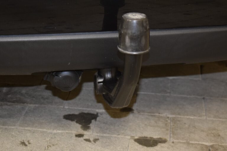 Close-up of a car's tow bar and electrical socket mounted to the rear bumper. The tow bar has a protective cap, and there are some oil stains on the ground beneath the vehicle.