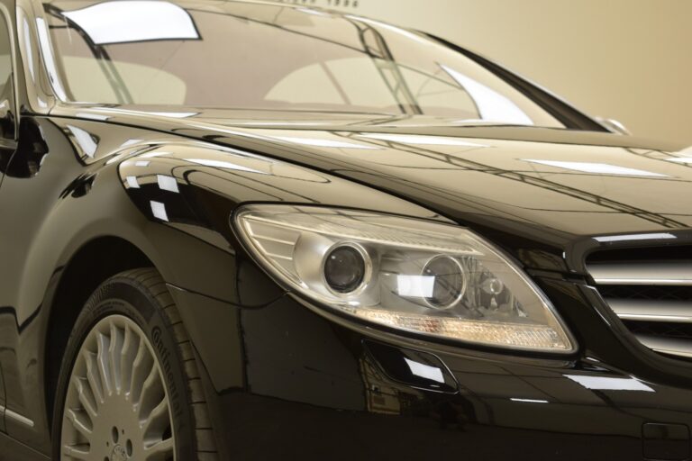 Close-up of the front of a black luxury car showing the headlight, grille, and part of a wheel, reflecting the well-lit environment.