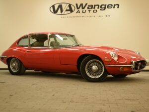 A classic red sports car with a sleek design is parked indoors on a tiled floor with the Wangen Auto logo on the wall behind it.