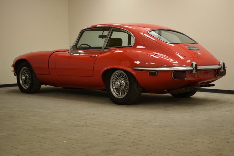 A red vintage car with a sloping back is parked in a beige-walled indoor space, viewed from the rear left side.