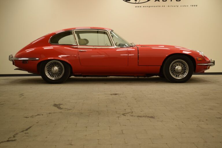 A classic red sports car with a sleek, rounded body design is parked indoors on a tiled floor against a beige wall.