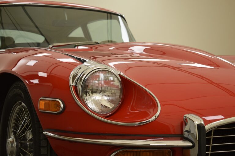 Close-up of the front of a red vintage car with a curved hood, round headlight, and chrome details. The background is a plain beige wall.