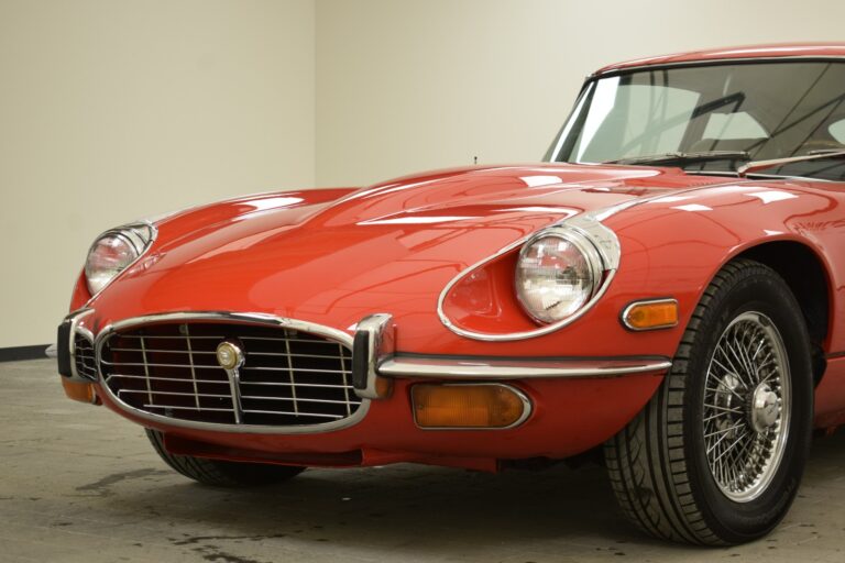 Close-up of a vintage red sports car with a sleek, rounded front design, featuring chrome accents and wire-spoked wheels. The car is parked indoors on a concrete floor.