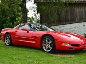 A red sports car is parked on a grassy area in front of a wooden building with trees in the background.