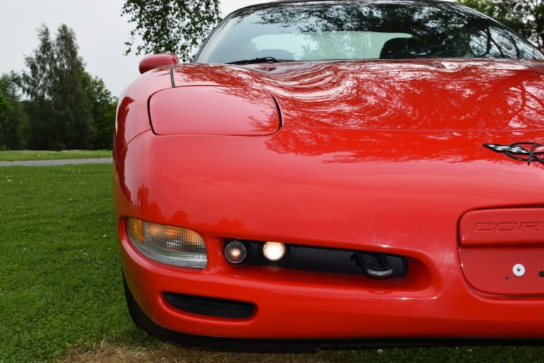 Red sports car, likely a Chevrolet Corvette, shown from the front, parked on grass with a tree and a blurred background.