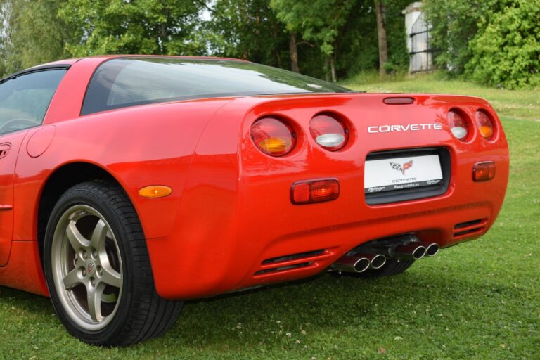 Rear view of a red Chevrolet Corvette with dual exhaust pipes, parked on a grassy area near trees and a building in the background.