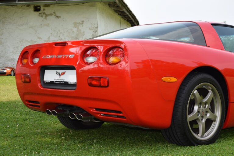 A red Chevrolet Corvette is parked on a grassy lawn. The image shows the rear end of the car, highlighting its taillights, dual exhaust pipes, and Corvette branding on the back.