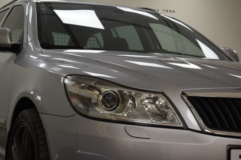 Close-up view of the front of a silver car, focusing on the right headlight and part of the grille. The indoor lighting creates reflections on the car's surface.