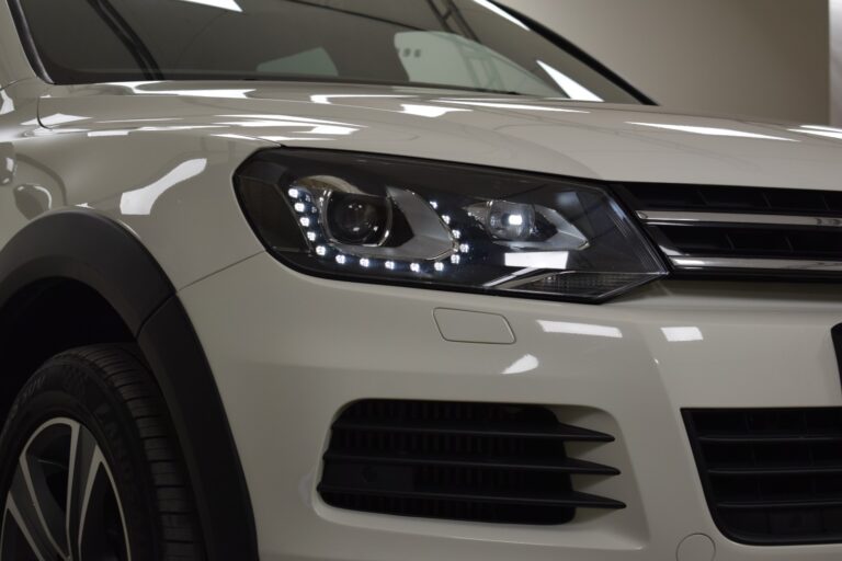 Close-up of the front right headlight and part of the front grille of a white SUV. The headlight is illuminated, showcasing LED daytime running lights.