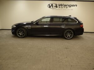 A dark-colored station wagon car is parked indoors on a tiled floor, with "Wangen Auto" signage on the wall in the background.