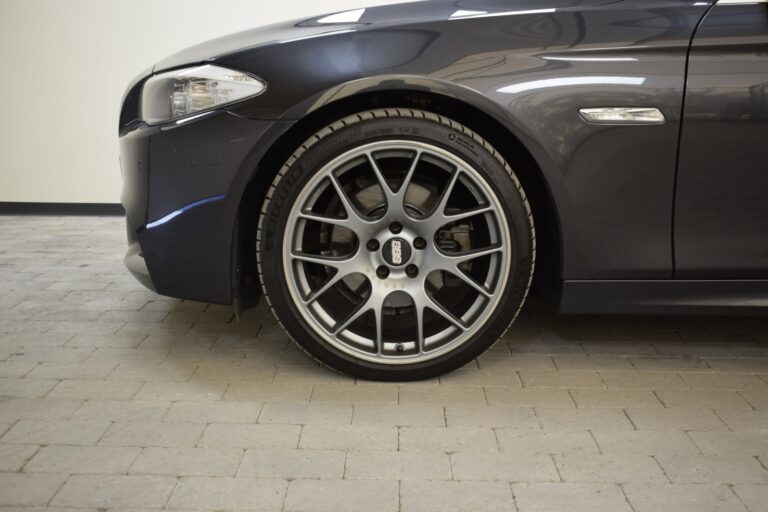 Close-up of the front left wheel and side mirror of a dark-colored luxury car parked on a tiled floor.