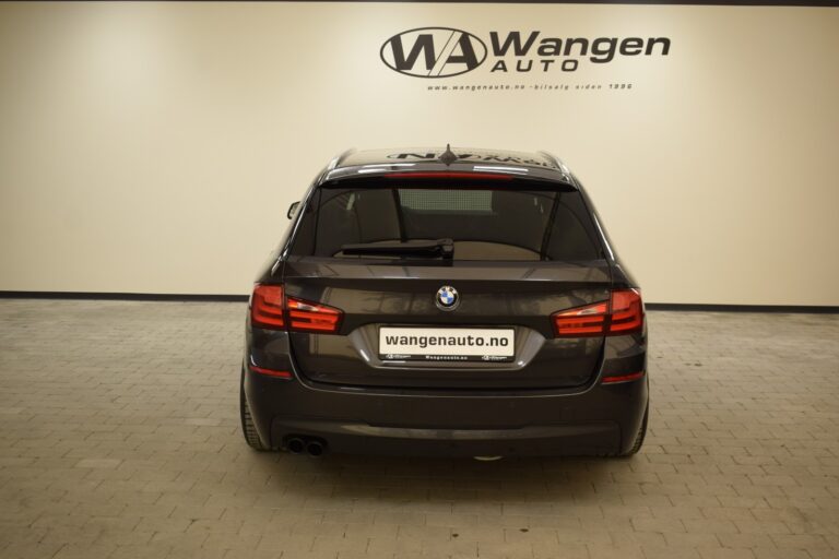 A black BMW car is shown from the rear, parked indoors against a beige wall with the Wangen Auto logo above it. The vehicle has a visible license plate with the dealership's name and website.