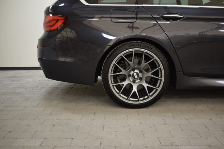 Close-up of the rear left side of a dark-colored car, highlighting the stylish alloy wheel and tire on a tiled floor in an indoor setting.