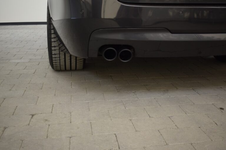 Close-up of the rear bumper and dual exhaust pipes of a dark-colored car, parked on a brick-paved surface.