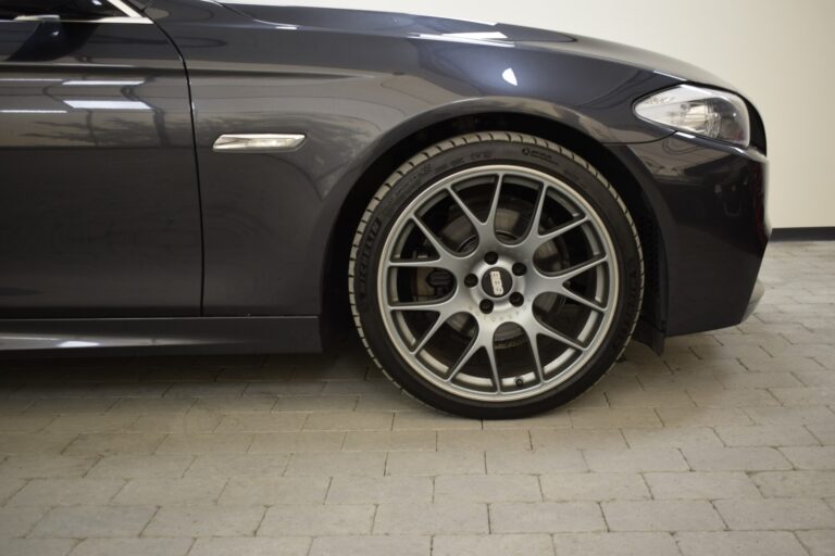 Close-up view of the front left side of a dark-colored car, focusing on the alloy wheel and tire, parked on light gray paving stones.