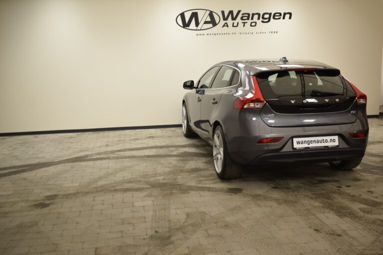 A grey hatchback car is parked in a showroom with a Wangen Auto sign on the wall. The car's license plate displays "wangenauto.no.