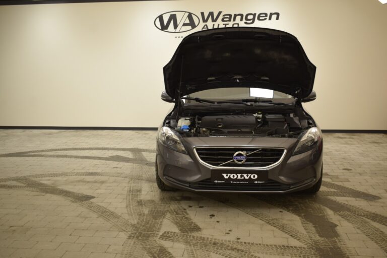 A gray Volvo car is parked indoors with its hood open. The wall behind it displays the "Wangen Auto" logo.