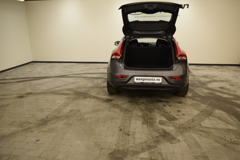 A gray car with its rear hatch open is parked on a tiled floor in an indoor setting. The car has a "wangenauto.no" license plate. Tire marks are visible on the floor.