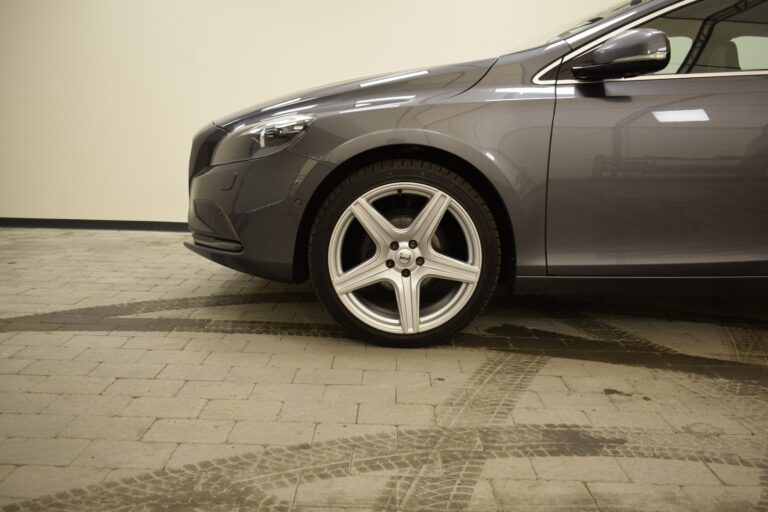 Front side view of a parked grey car on a tiled surface, focusing on the front wheel and tire. The background is a plain, off-white wall.