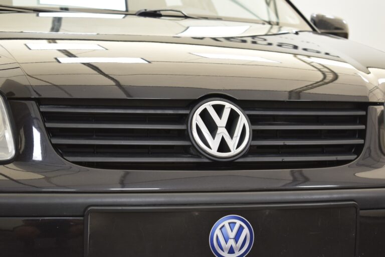 Close-up of the front grille and logo of a black Volkswagen car, with the VW emblem clearly visible in the center. The car's glossy finish reflects the surrounding lights and environment.