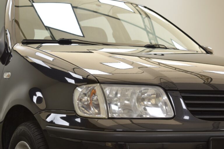 Close-up view of the front end of a black car, highlighting the headlight and part of the hood under bright indoor lighting.