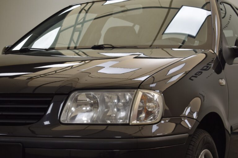 Close-up of the front left side of a black car showing the headlight, hood, and part of the windshield in an indoor setting with reflections of ceiling lights.