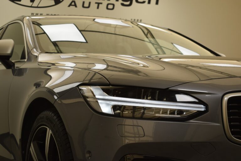 Close-up of the front of a silver car showcasing its left headlight and part of the grille, with a reflection of the ceiling lights on the hood.