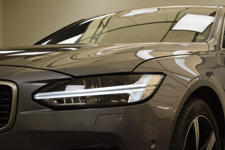 Close-up of a sleek, modern car with a focus on the front left headlight and part of the hood and windshield. The car is metallic gray with illuminated headlights.