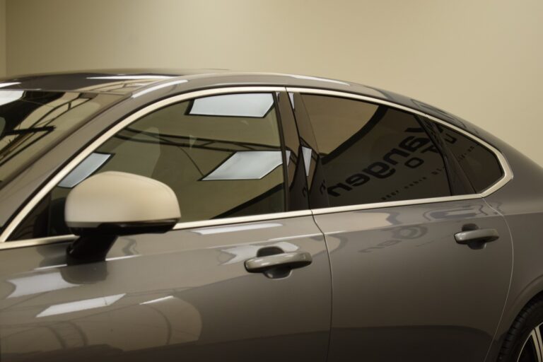 Side view of a modern gray car with tinted windows, showing part of the door and rearview mirror. The reflection of ceiling lights is visible on the car roof.