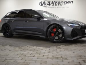 A gray Audi RS6 Avant with black and red wheels is parked in an indoor showroom with "Wangen Auto" signage on the wall behind it.