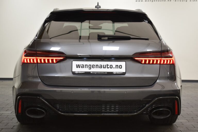 Rear view of a gray car with illuminated tail lights, dual exhausts, and a license plate displaying "wangenauto.no". The car is parked indoors against a light-colored wall.