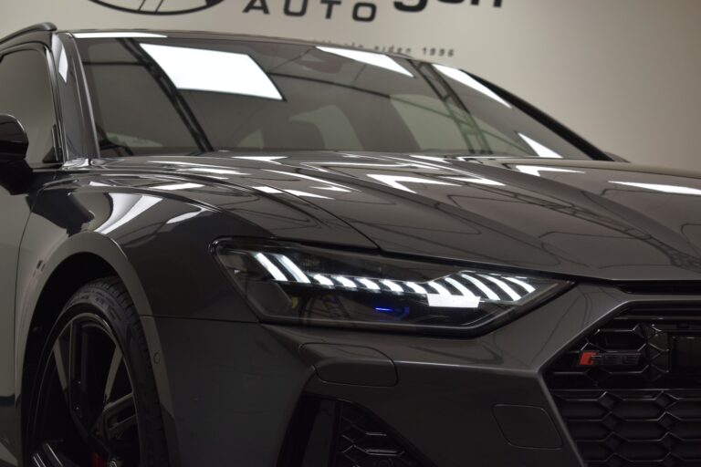 Close-up of a sleek, dark-colored car with illuminated headlight, showcasing modern design and detailing. Part of car’s logo and a portion of background wall signage are visible.