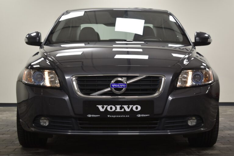 A front view of a dark-colored Volvo car with illuminated headlights, displayed in an indoor showroom setting.