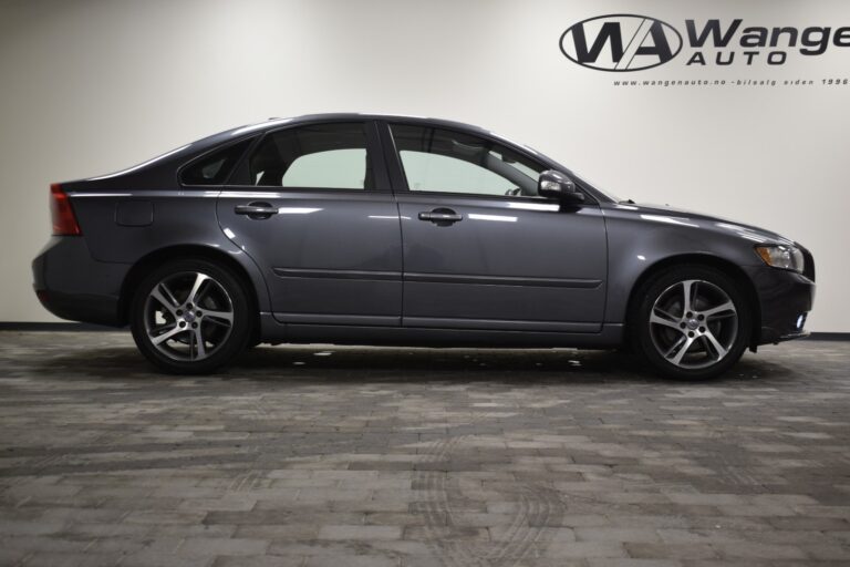 A side view of a dark gray Volvo sedan parked on a stone-tiled floor, with the logo and text "Wanger Auto" visible on the wall in the background.