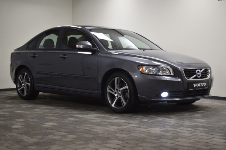 A gray Volvo sedan is parked in an indoor garage with a Volvo sign visible on the front. The car is viewed from a slight angle, showing both its front and side.