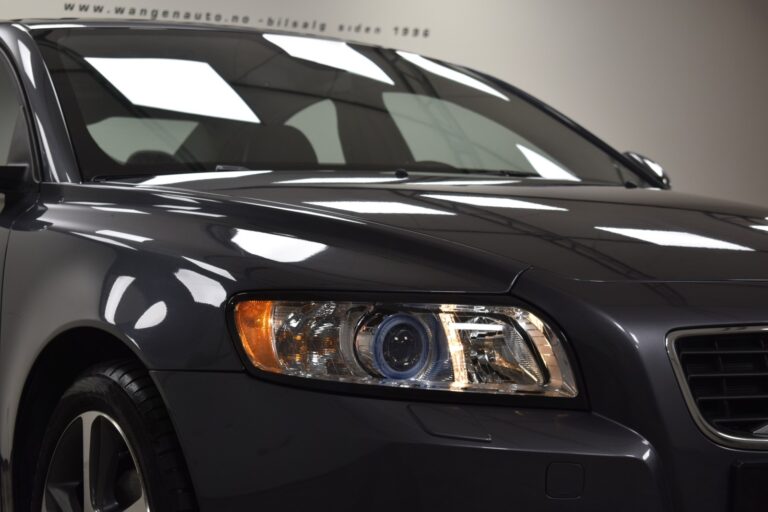 Close-up of the front left side of a dark grey car showcasing the headlight, part of the grille, and the side mirror. The car appears to be displayed indoors under bright lighting.