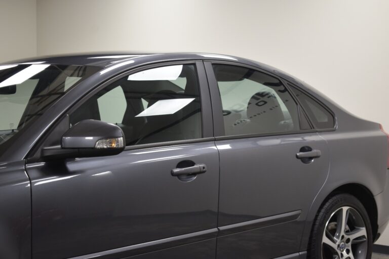 A gray sedan's side view, showing the driver's side mirror, front door, and rear door with tinted windows under indoor lighting.