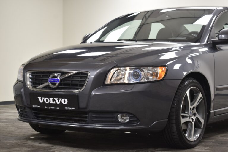 Close-up of a gray Volvo sedan front view showing headlights, grille with the Volvo logo, and part of the windshield, parked indoors.